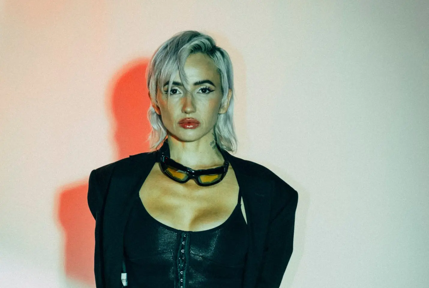 Lights with sunglasses aroud her neck and black top on posing against a plin wall