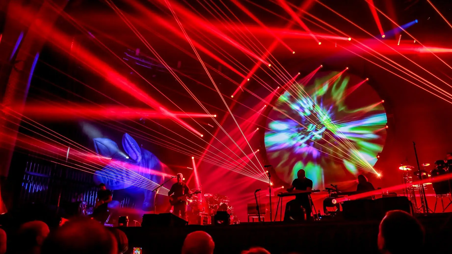 Brit Floyd speelt in silhouet op een groots verlicht podium, omringd door felle rode laserstralen en een groot rond scherm met kleurrijke psychedelische projecties.