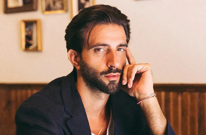 Portret van influencer Davide De Pierro met donker haar en baard, zittend in een café. Hij draagt een donker jasje en leunt met zijn hand tegen zijn gezicht. Op de achtergrond hangen ingelijste foto’s aan de muur.