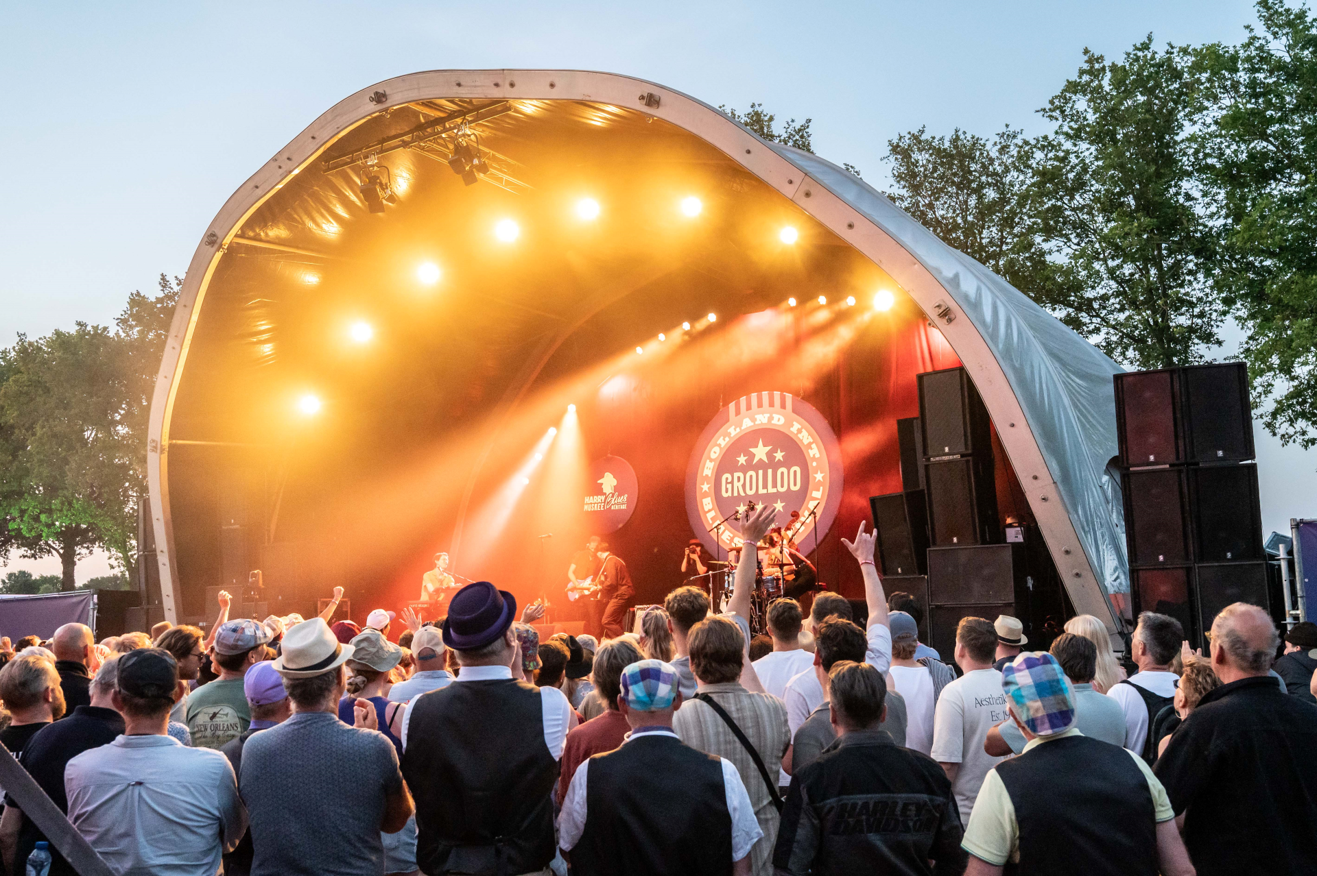 Een zomerse avond op het **Holland International Blues Festival** in Grolloo, waar een enthousiast publiek geniet van live bluesmuziek onder warme podiumlichten.