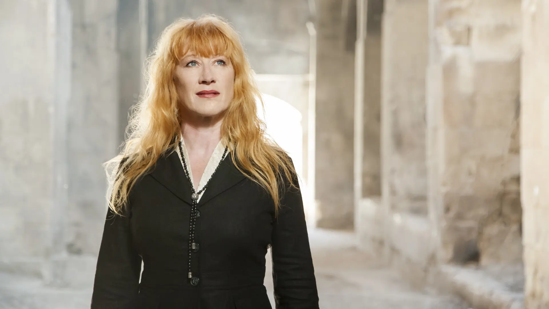 Loreena McKennitt met lang rood haar en een zwarte jas, staand in een stenen gang met licht op de achtergrond.