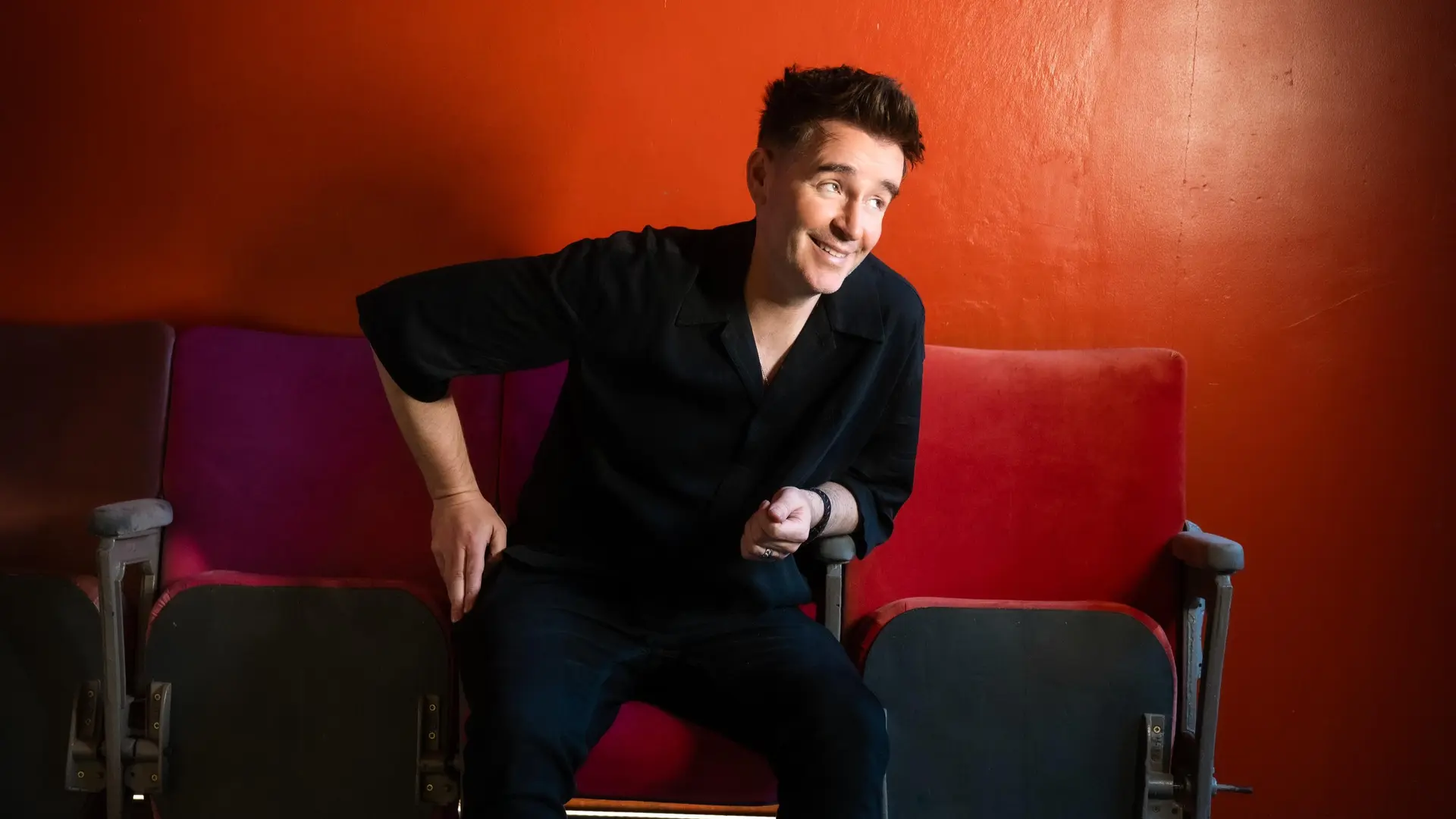 Jarlath Regan zit op rode theaterstoelen voor een rood-oranje muur, draagt een zwart overhemd, donkere broek en bruine laarzen. Op de vloer naast hem ligt een microfoon met kabel.