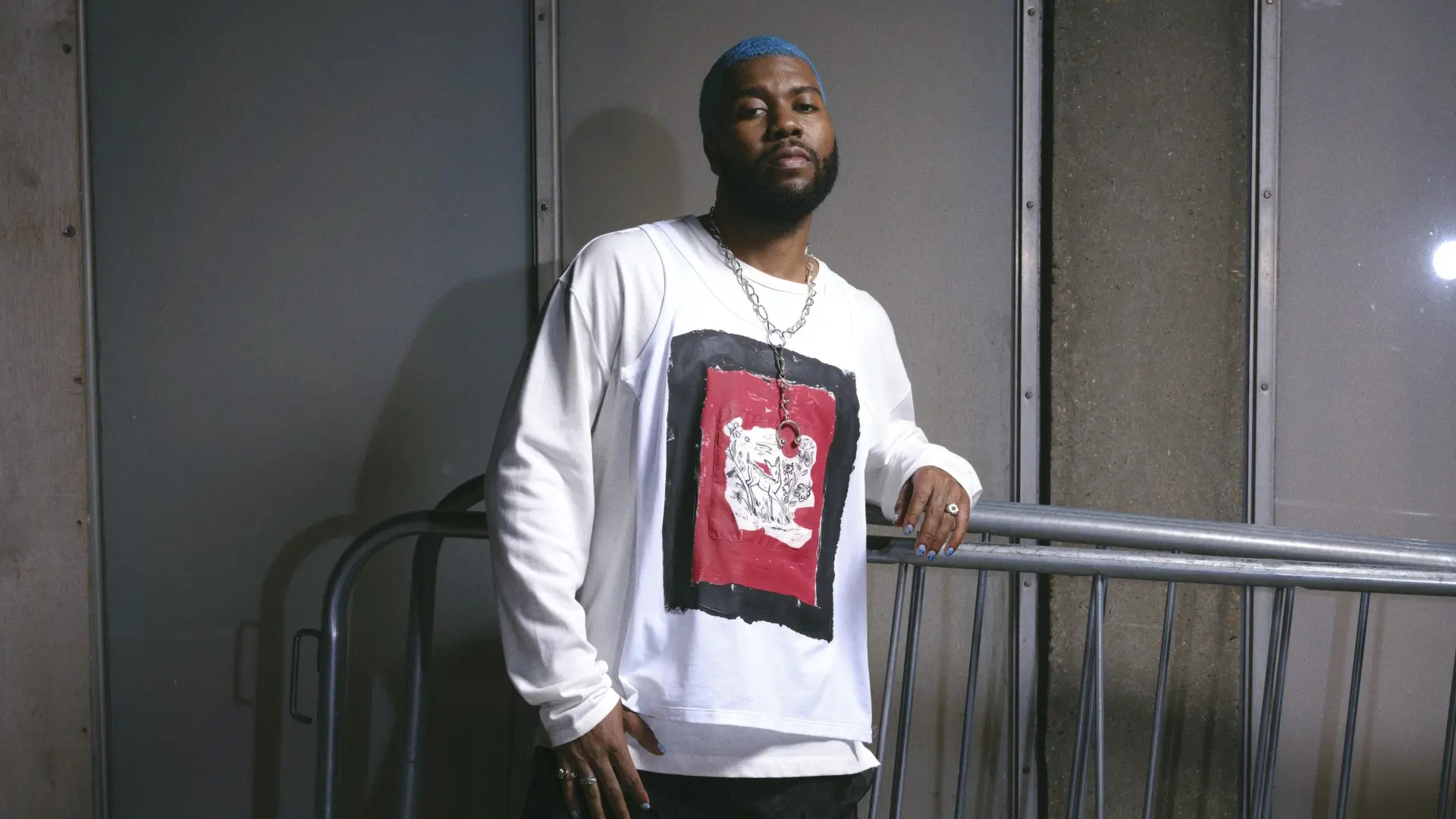 Khalid staat leunend tegen een metalen railing, draagt een wit shirt met rode en zwarte print, zwarte broek en zilveren ketting, met blauw geverfd haar in een industriële omgeving.