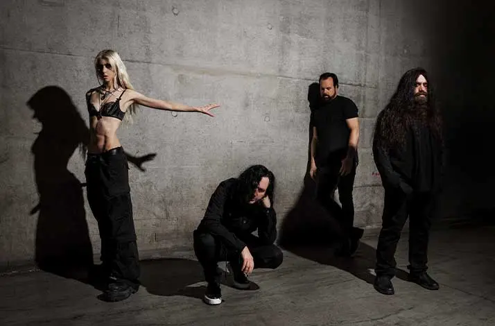 Vier bandleden van The Pretty Reckless tegen een betonnen muur: links een vrouw met lang blond haar in zwarte bh en zwarte broek met arm gestrekt opzij, daarnaast een man gehurkt in zwart, rechts twee staande mannen in zwart.