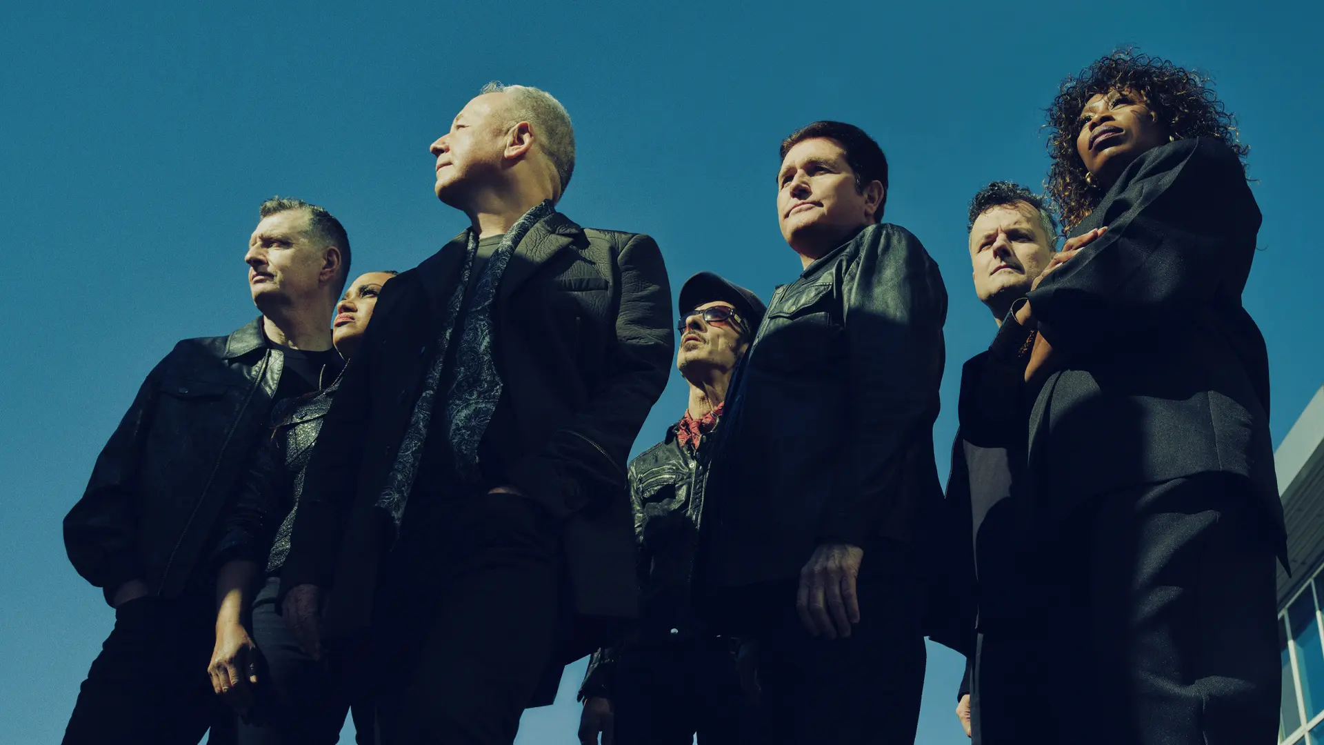 Simple Minds poseert met zeven bandleden in donkere kleding, van onderaf gefotografeerd tegen een strakblauwe lucht.