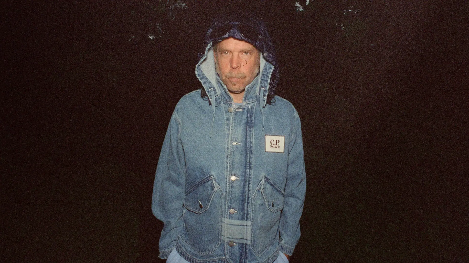 Bonnie ‘Prince’ Billy staat ’s nachts buiten, draagt een blauwe spijkerjas met capuchon en een patch met tekst “C.P. Palace”, handen in de zakken, donkere achtergrond met bomen