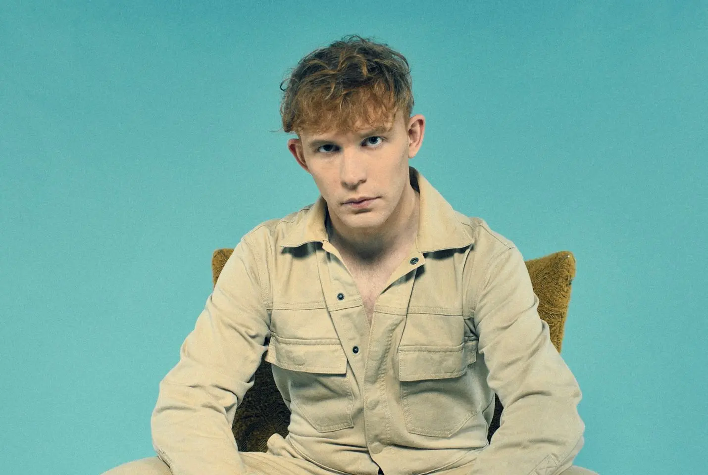 A man in a beige jacket sat in front of a blue backdrop