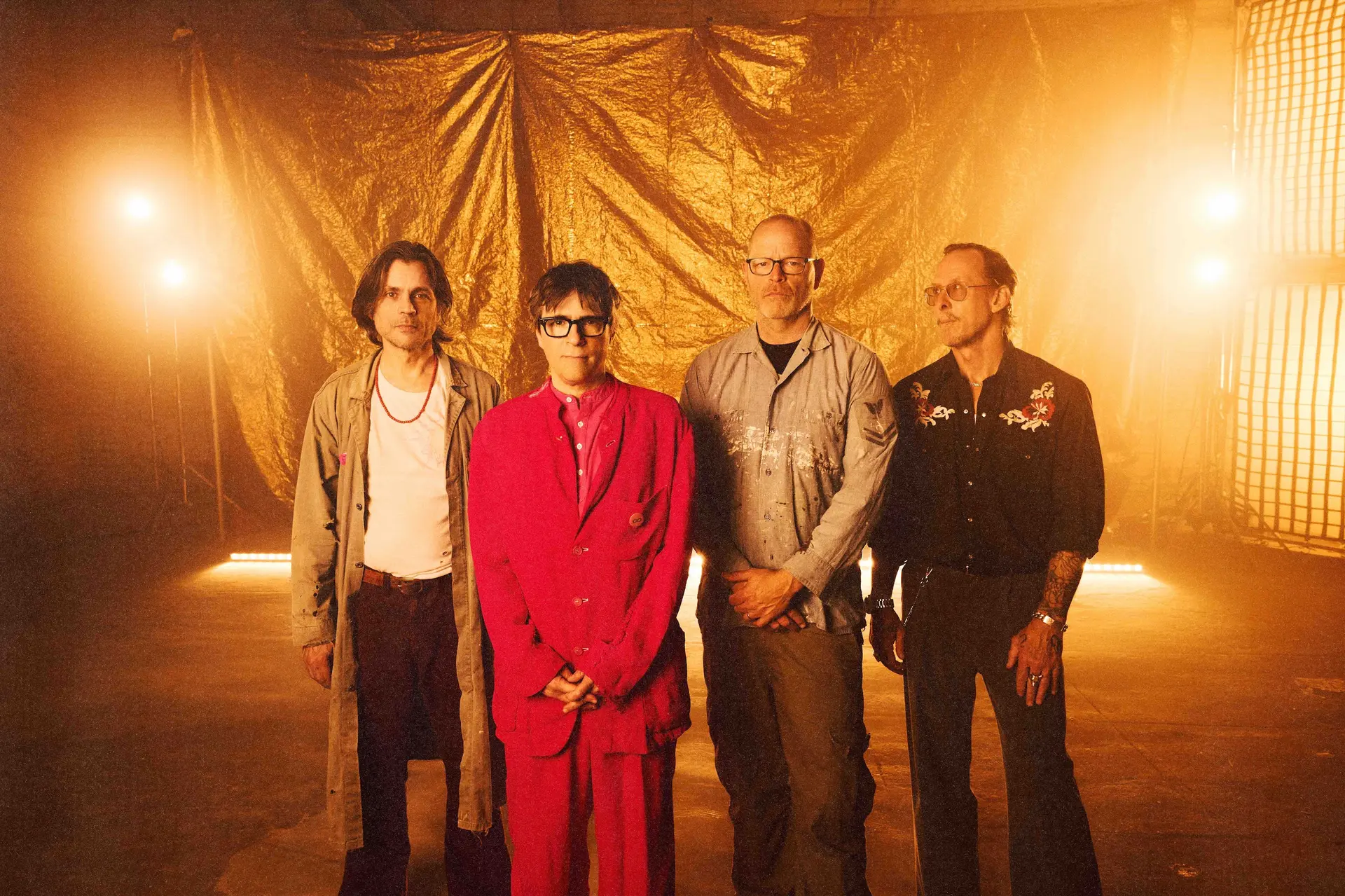 Vier leden van de rockband Weezer poseren naast elkaar in een industriële ruimte met warm oranje licht. Frontman Rivers Cuomo draagt een felrood pak, terwijl de andere bandleden in casual kleding voor een goudkleurige achtergrond staan.