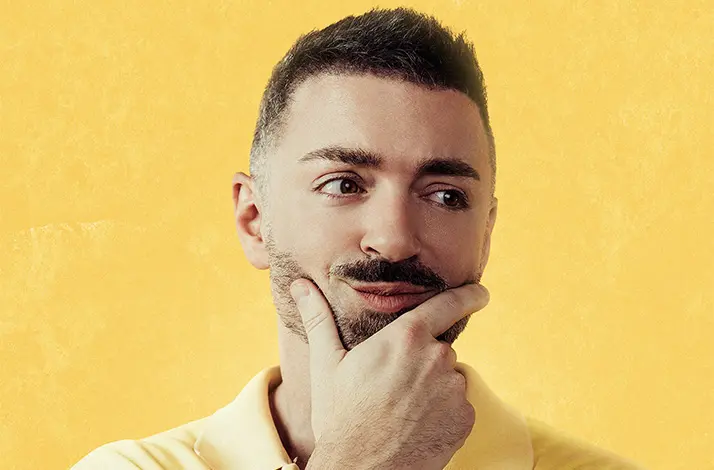 Matteo Lane voor een gele achtergrond. Hij draagt een geel shirt.