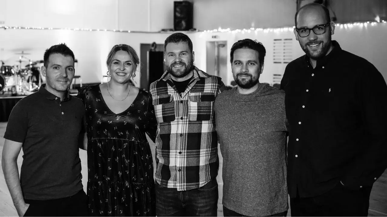 Black and white photo of five people standing close together indoors, smiling at the camera. They are in a casual setting with string lights in the background and music equipment visible on the left.