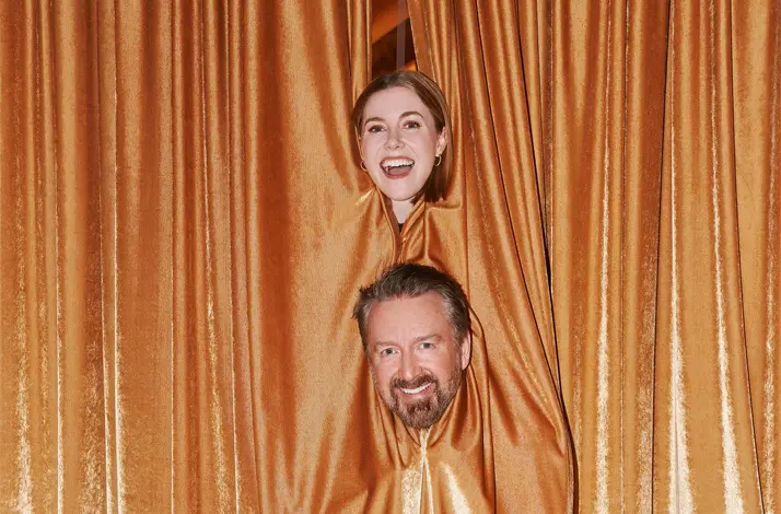 Chad Daniels en Kelsey Cook steken hun hoofd door openingen in gouden theatergordijnen. Ze lachen naar de camera. De achtergrond bestaat uit glanzende gordijnen en een rood tapijt, wat een speelse, theatrale sfeer geeft.