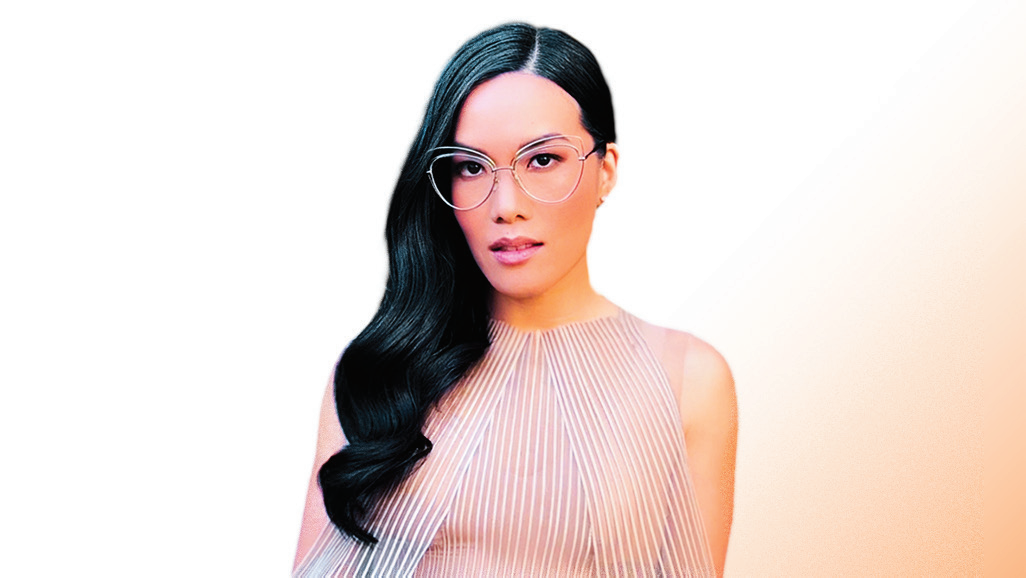 Portret van Ali Wong met lange zwarte haren over één schouder, bril met dun montuur en een lichtroze geplooide mouwloze top, tegen een lichte egale achtergrond.