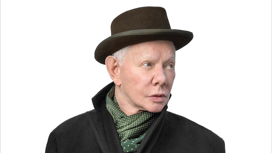 De Britse muzikant Joe Jackson, gekleed in een donkere jas en bruine hoed met een groene sjaal met stippen, houdt een porseleinen theekopje vast en kijkt naar rechts. De achtergrond is wit.