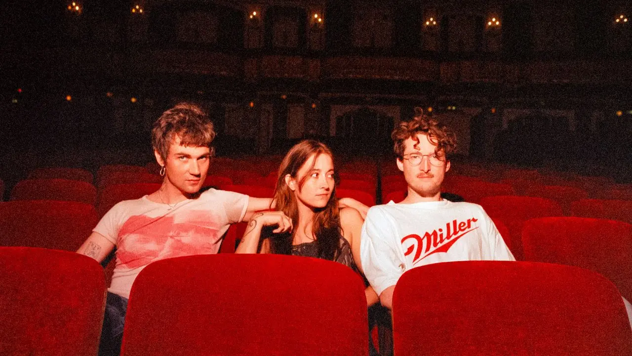 Three people sit in red theater seats, with the two on the sides leaning toward the person in the middle.