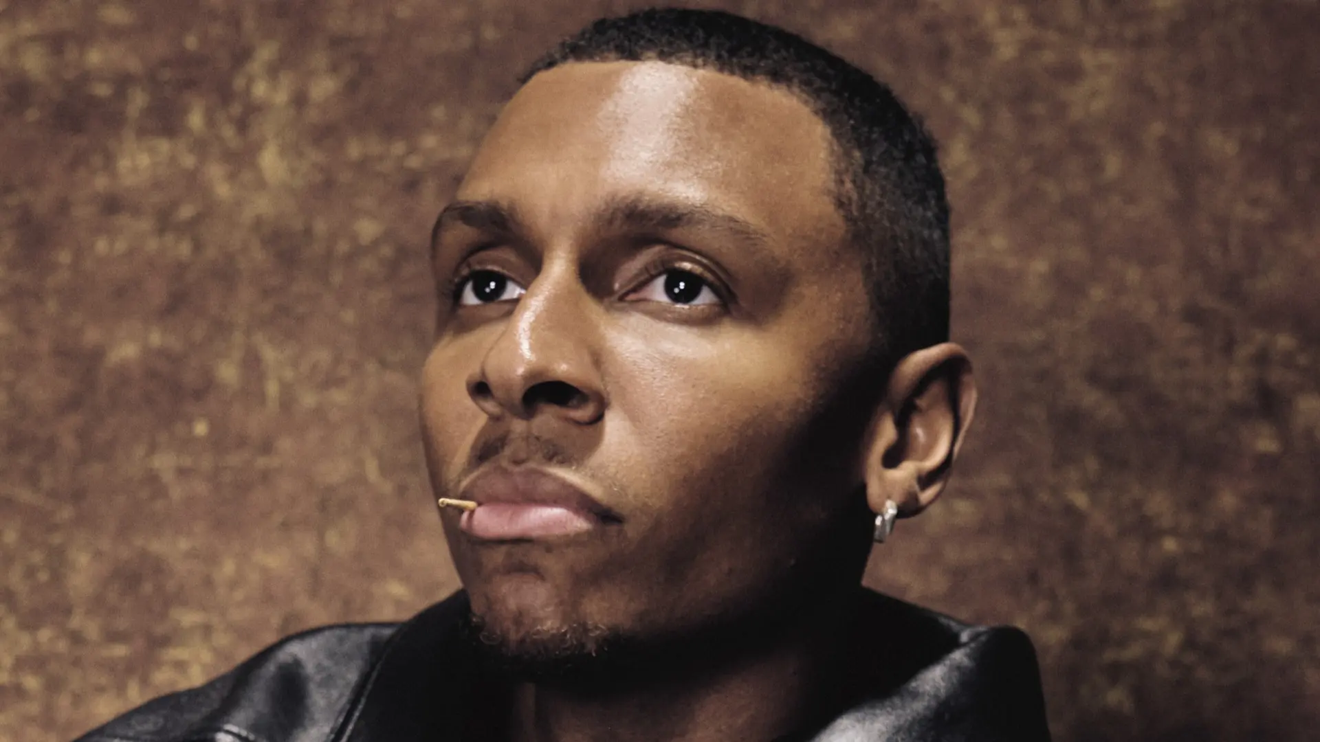 Portret van Masego in een zwarte leren jas die peinzend omhoog kijkt tegen een warme bruine achtergrond.