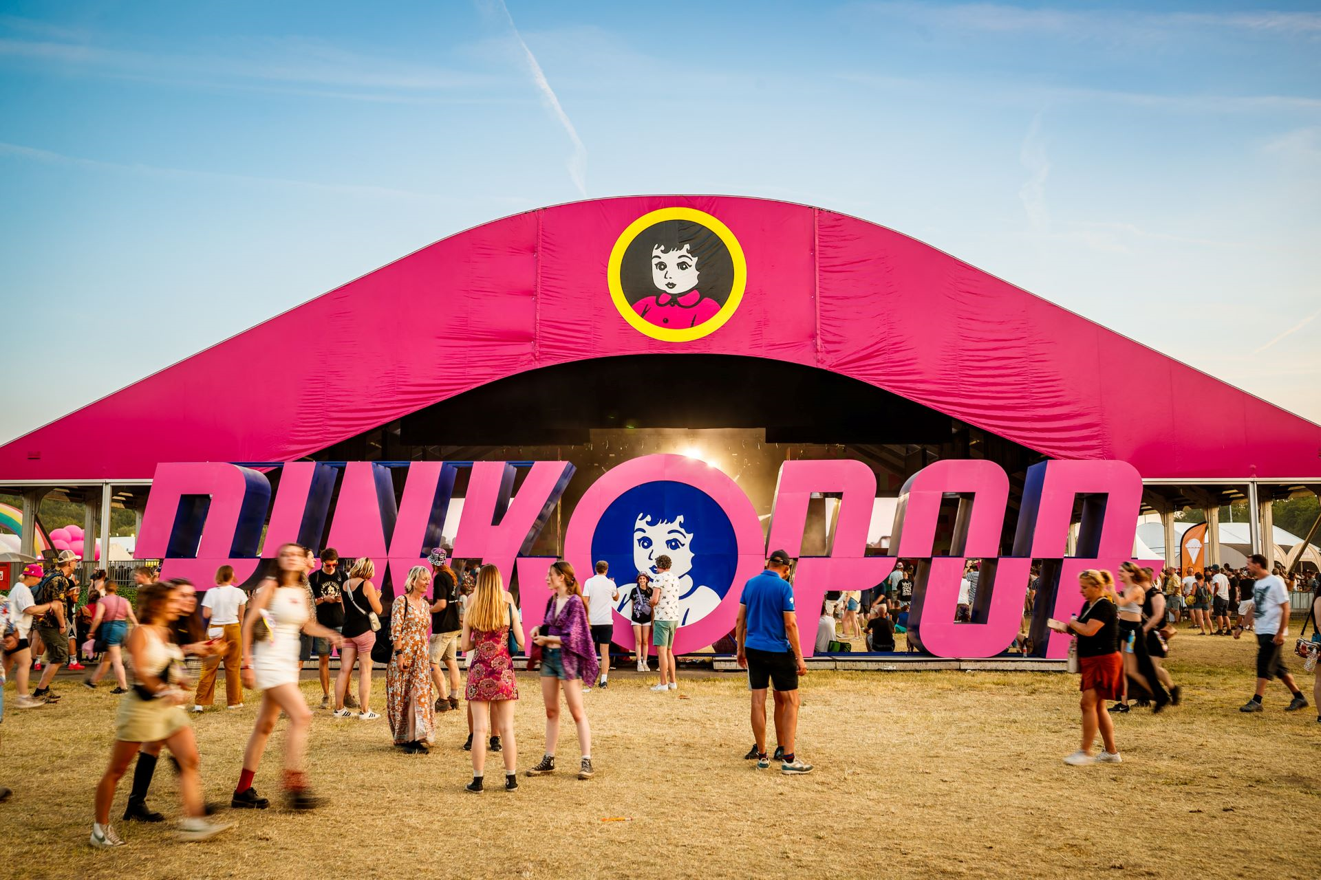Een zonnige dag op Pinkpop, waar festivalgangers rond de iconische roze tent en het grote logo samenkomen voor muziek en sfeer.