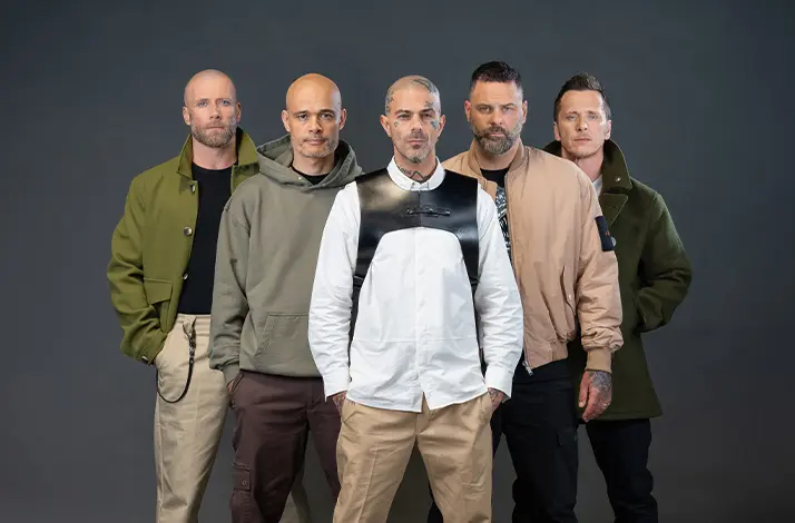 De vijf bandleden van de boyband Five staan naast elkaar in een studio tegen een donkere achtergrond, casual gekleed in jassen en sneakers, met serieuze blik en zelfverzekerde houding.
