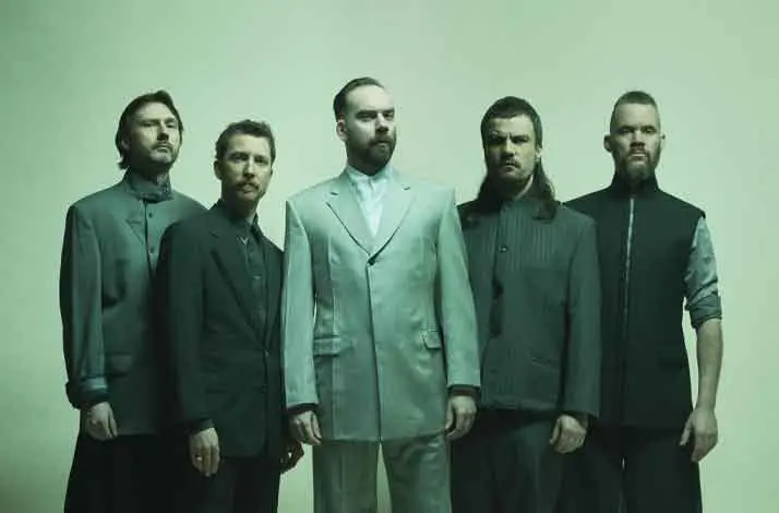 Vijf bandleden van De Staat staan naast elkaar in minimalistische, donkere outfits tegen een groenige achtergrond, met serieuze blikken en een strakke, gestileerde, bijna futuristische uitstraling.