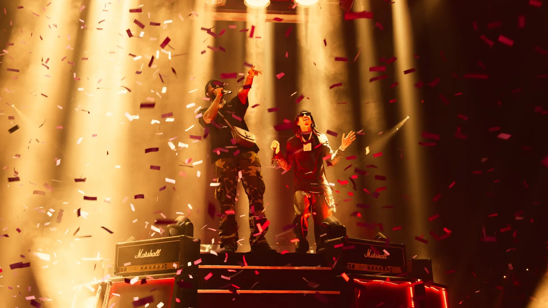 D-Block Europe staat centraal op een verhoogd podium tussen dwarrelende rode en witte confetti, gehuld in zwart en camouflage, terwijl krachtige goudgele lichtbundels van boven een uitbundige, warme concertsfeer creëren.