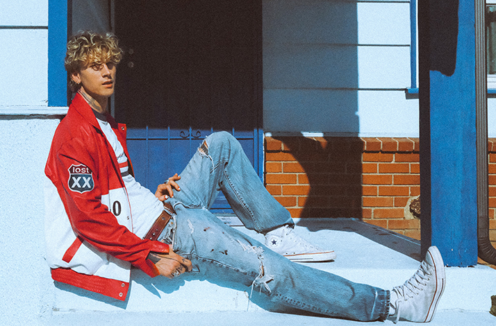 Machine Gun Kelly sitting on a patio, wearing a red leather jacket, blue ripped jeans and white sneakers