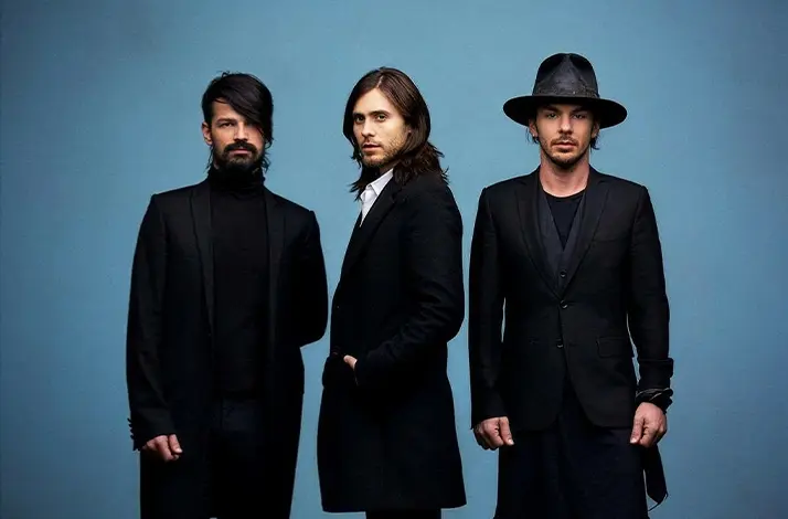 Drie leden van Thirty Seconds to Mars staan tegen een effen blauwe achtergrond: links gitarist Tomo Miličević, in het midden zanger Jared Leto en rechts drummer Shannon Leto. Ze dragen donkere, stijlvolle kleding en kijken serieus recht in de camera.