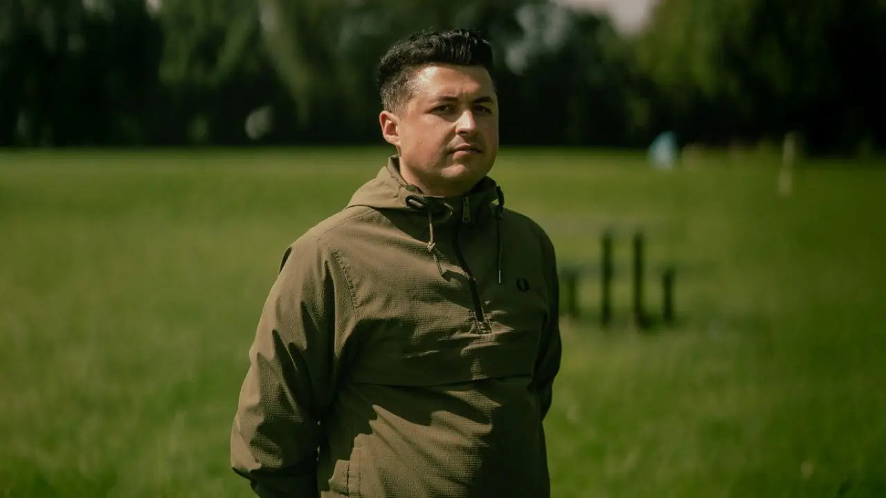 A man with dark hair in a green coat stands in a field