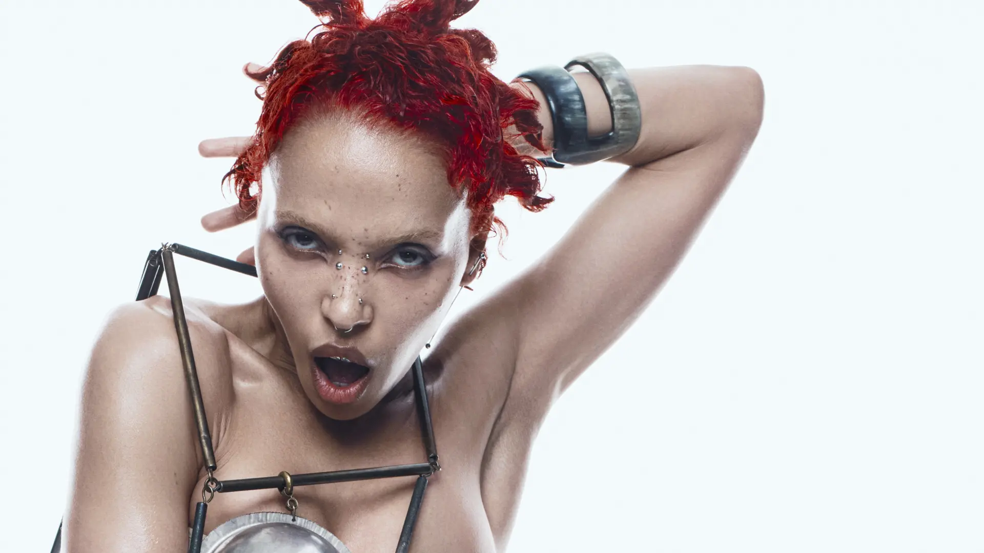 FKA Twigs met rood krullend haar, zichtbare sproeten en meerdere gezichtspiercings, met ontbloot bovenlichaam en een metalen harnas om de schouders en borst, één arm achter het hoofd en open mond, tegen een witte achtergrond.