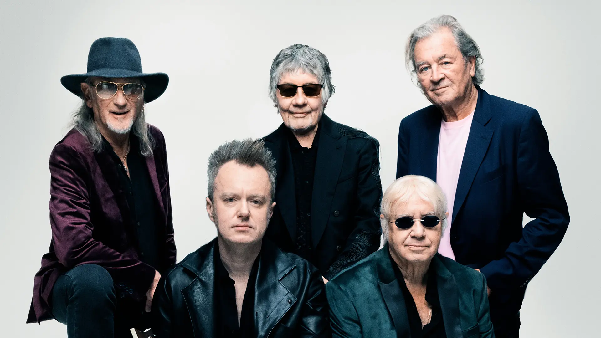 Groepsportret van Deep Purple met vijf leden. Ze poseren voor een lichte achtergrond, dragen donkere, stijlvolle kleding en sommige leden hebben zonnebrillen op. Eén persoon links draagt een brede hoed, twee leden zitten vooraan en drie staan achter hen.