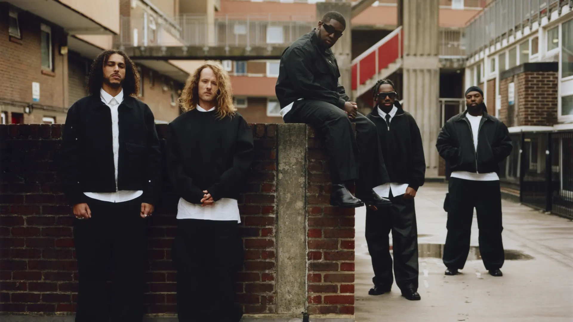 De band Ezra Collective poseert op een betonnen galerij tussen flatgebouwen, met vijf bandleden verspreid over de ruimte.