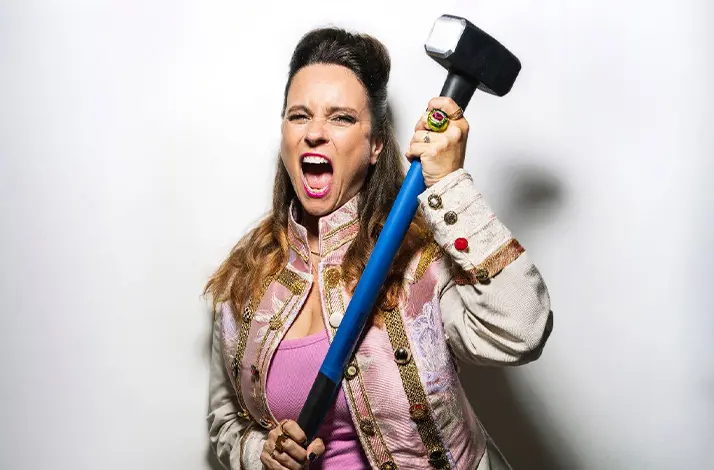 Tova Leigh staat tegen een witte achtergrond en houdt een grote voorhamer omhoog. Ze draagt een lichtroze top met een versierde jas en zwarte broek, en kijkt met open mond krachtig en energiek in de camera.