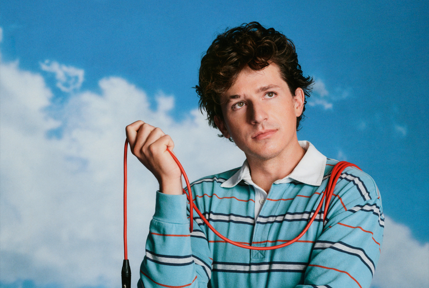 Charlie puth in striped top, sky in background, holding a microphone with the wire over his shoulder