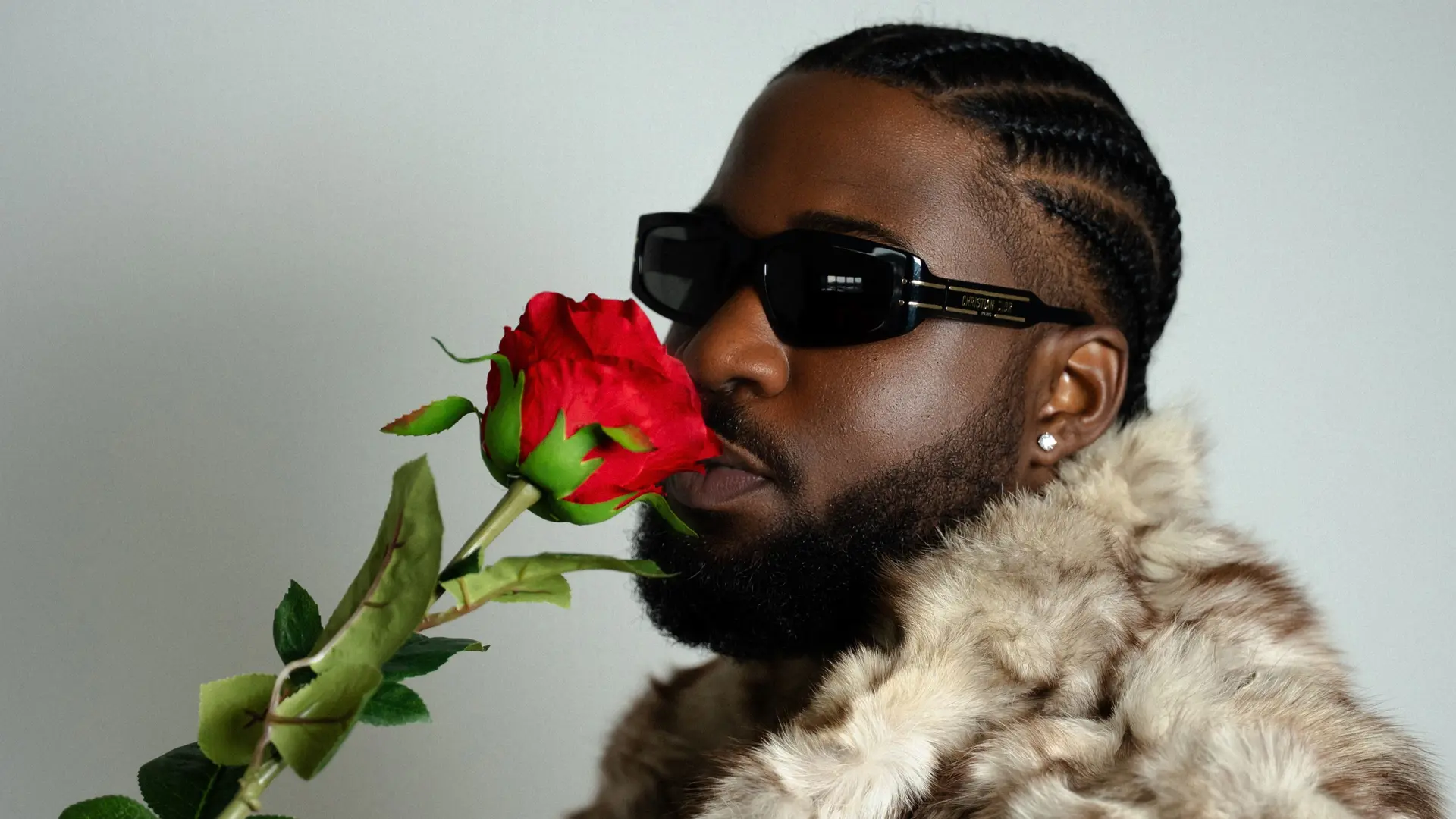 Man met ingevlochten haar en zonnebril die een rode roos vasthoudt en ruikt, gekleed in een bontjas.