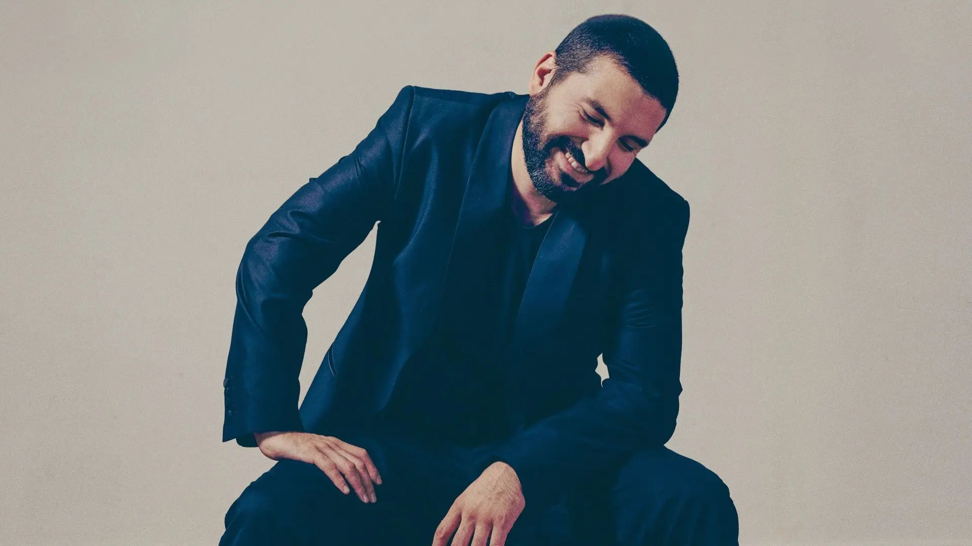 Ibrahim Maalouf zittend, gekleed in een donker pak met witte sneakers, licht voorovergebogen en glimlachend.